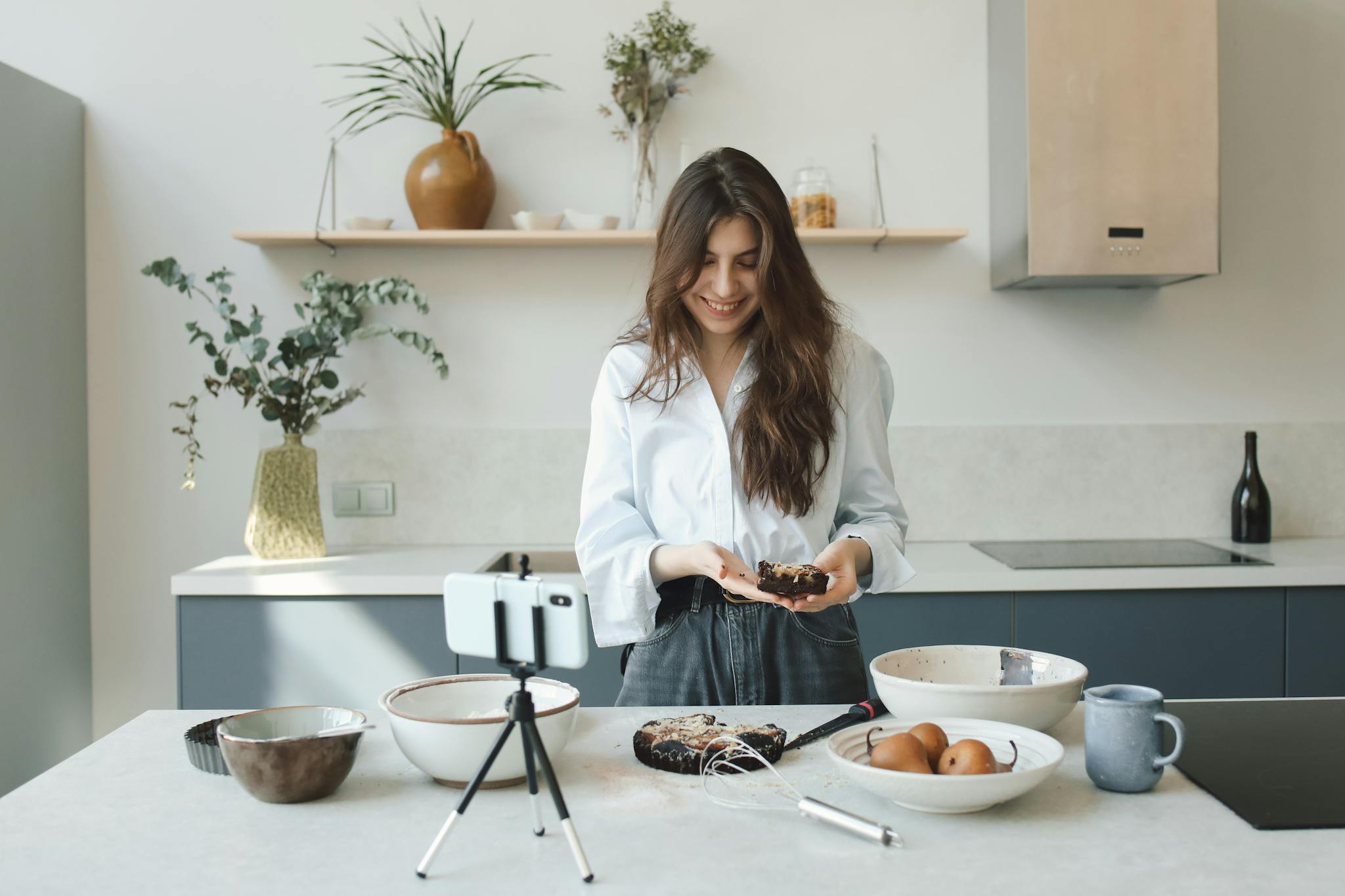 A woman smiles while baking and filming a video in a modern kitchen.