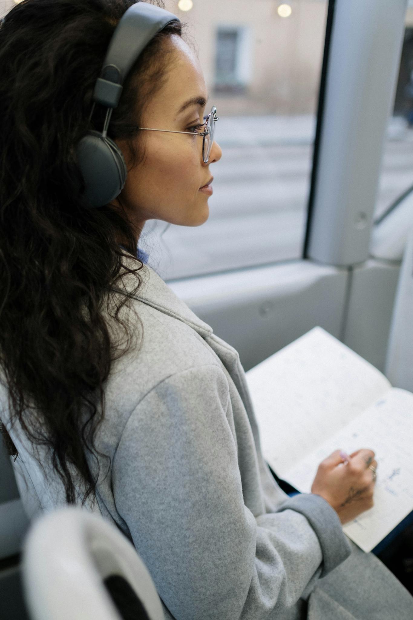 Side view of a woman with headphones writing in a notebook while sitting on a bus.